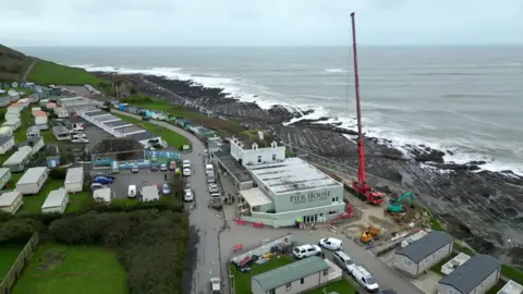 An aerial view of The Pier House restaurant and bar overlooking the sea at Westward Ho! with a large red crane to the right of it. There are static holiday homes surrounding the restaurant.
