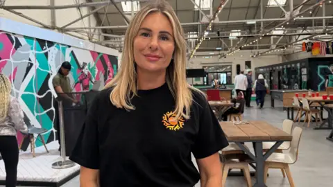 A woman with long blonde hair, wearing a black shirt with a yellow and orange logo, smiles as she stands in a large hall as an event takes place.