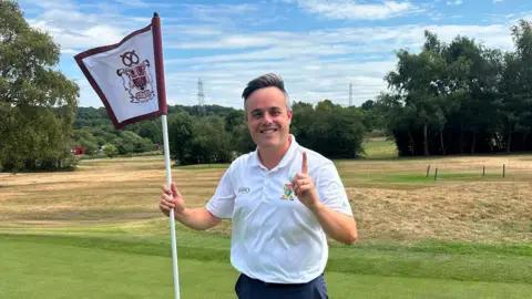 Matthew Vigurs holding up a white a burgundy flag on a golf course while holding up his other hand and pointing upwards to signify the number one. He is wearing a white polo top and has swept back dark hair.