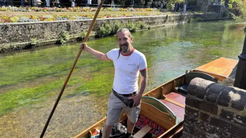 Jo Burn/BBC A man in a white t-shirt and shorts stood in a wooden boat, which is floating in clear, shallow water. He is holding a long stick, which he is using to keep the boat stable.