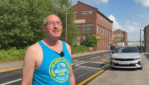 A man wearing a blue vest top with a yellow logo in the middle of it. He is smiling, looking slightly off to the right of the camera. There is a tall brick building in the background behind him and a silver car to the side of him.