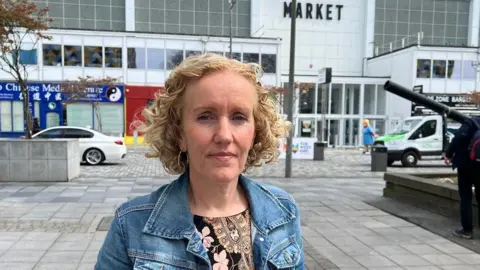Victoria Allen is wearing a pink and black patterned top and a blue denim jacket and is standing in a shopping street in Plymouth with a sign for the market in the background