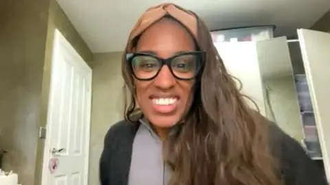 Jhiselle Feanny smiling at the camera in a room with a brown head band over her dark brown hair, wearing thick rimmed dark glasses