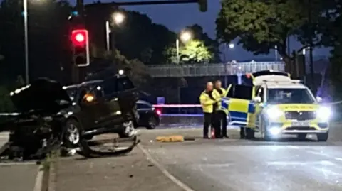 The scene of the crash. A black vehicle is off the road to the left and has damage, while two police officers are stood next to a marked police vehicle.