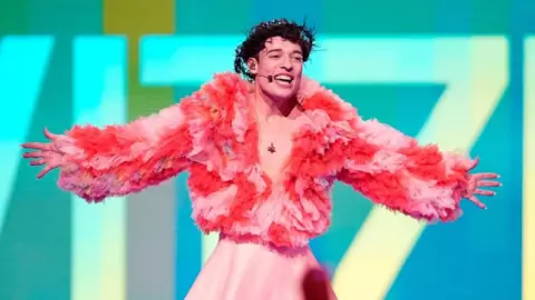 Getty Images Nemo with short brown hair, wearing a pink dress and red and pink puffy jacket, performing at Eurovision