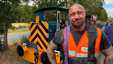 Lambert stands with his hands on his hips and is smiling. He wears a black harness over a high vis vest. A herritage locomotive with yellow and black stripes on the front is on the track behind him.