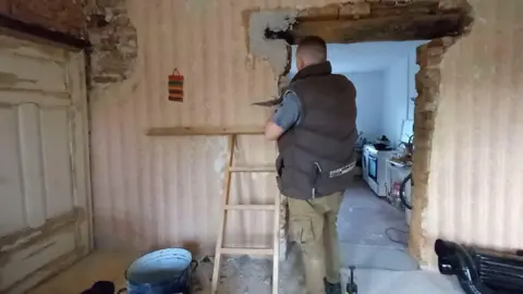 Jordan Vye A video still of Lewis Vye plastering around a doorway inside the house. His back is to the camera and he's wearing green cargo trousers and a padded bodywarmer. The walls are a mixture of old pink wallpaper and exposed brick.