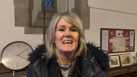 A woman with dark blonde hair wearing a black and white top with a black puffer coat inside a church