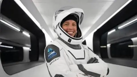 SpaceX/O2XR A woman in a white astronaut suit and helmet smiles at the camera with her arms crossed over her chest. The helmet is up so you can see her face. She stands in a white corridor with black windows.
