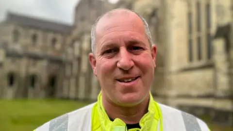 Jon is wearing a high-visibility firefighters jacket and standing in front of the cathedral and its grounds, which are out of focus. 
