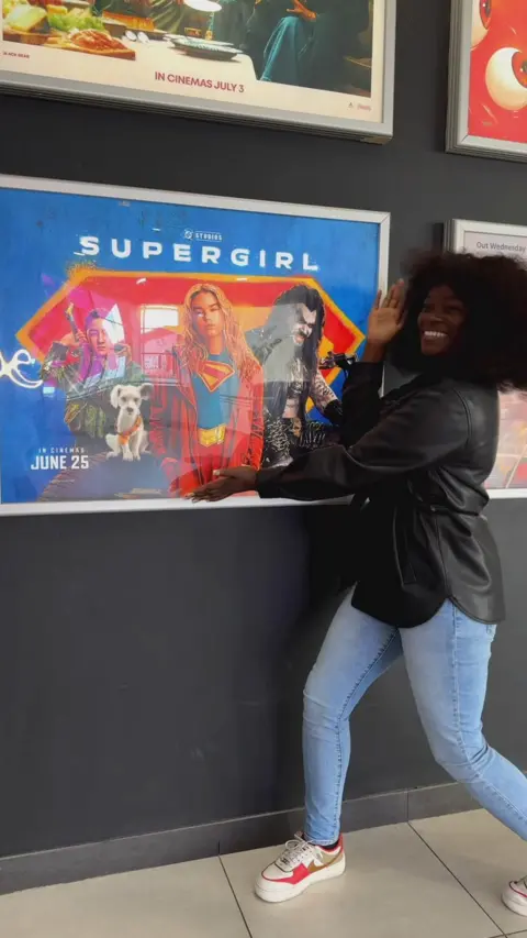 A woman in a black leather jacket is smiling at the camera. She is standing next to a Supergirl film poster