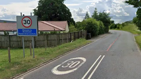 A road between fields with green grass with a 30mph road sign