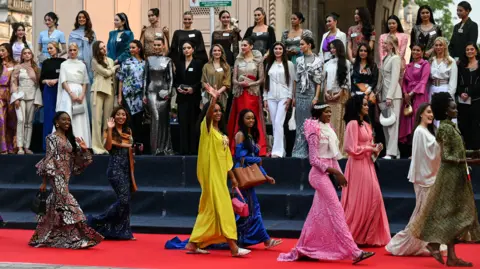 Noah Seelam / Getty Images Two rows of young women with name badges on a tiered podium with eight women walking in front of them on a red carpet in pairs - one of the women in a long yellow dress is waving