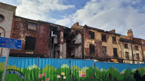 A number of crumbling old buildings some without windows or walls behind a series of metal hoardings painting blue with flower landscapes on them