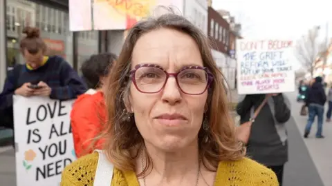 Qays Najm/BBC Sarah Fitch with long hair, wearing glasses and a yellow top. She is standing in front of people holding signs.