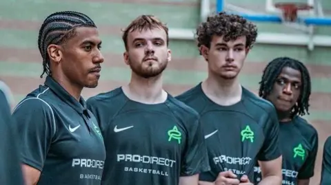 CourtesyOfZed Four men in green PRO:DIRECT Basketball T-shirts. Andre Arissol on the far left has short dark hair and beard. They are standing in front of a basketball hoop.