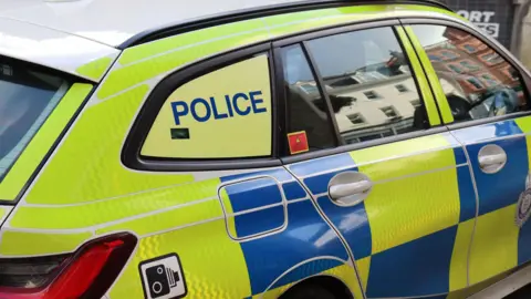 BBC The side of a police car. It has reflective blue and yellow checks, the lettering POLICE on the back window and a sticker with the picture of a speed camera on it. Buildings are reflected in its windows.