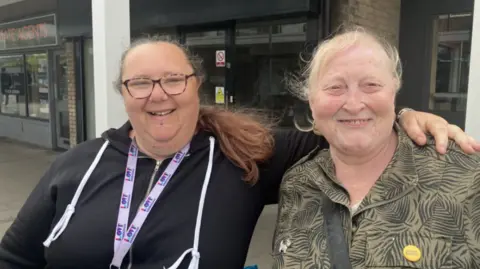 BBC/ Lara King Two women side by side. The woman on the left has dark hair and is wearing a black hoodie and has glasses on and she has her arm around the other woman who is wearing a khaki coloured top with a grey flower pattern. 
