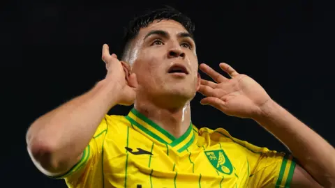 PA Media Marcelino Nunez cups his ears while wearing a yellow and green Norwich City top at night.