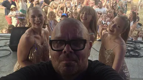 A selfie taken by Martin Cosser, who is wearing a black top and black-rimmed glasses. He is standing on a stage in front of three smiling women in sparkly dresses. They all have their backs turned to the crowd who can be seen cheering and smiling behind them