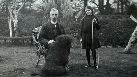 Michelle Bullivant A black and white image of William and Elsa Hall in a garden. He has a high white collar under a dark jacket and waistcoat and is sitting in a garden chair with a large woolly black dog in front of him. She has long dark hair pulled back from her face and is wearing a dark jumper over a dark skirt and is holding a hoop in her left hand.