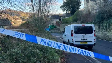 A scene showing blue and white police tape across a road. In front of the tape is a white van, the road is seen on the right hand side and greenery and a wall and part of a house can be seen on the left. In the distance the fire damaged house can be seen and beyond that more houses rising up a hill.