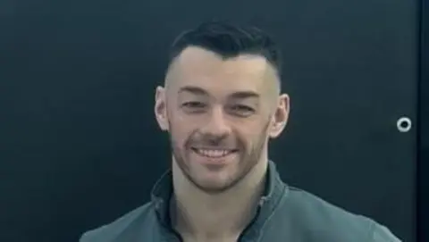 Josh Irwin Josh Irwin is standing behind a black backdrop and is smiling.
He has short black hair and is wearing a grey jacket and black T-shirt.