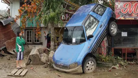 A young woman in a green and white jacket looks on forlornly as a blue car hangs from a tree, with its rear end in the air, having been lifted by floods during Typhoon Rai in the Philippines in 2021