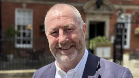 A man wearing a white shirt and blue jacket. He is looking directly at the camera and smiling for the photograph. There is a brick building with black railings behind him.