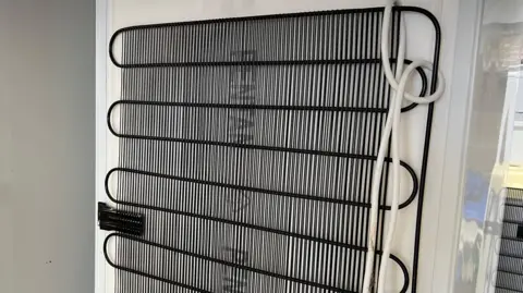 The back of a fridge showing the black coils and vertical strips of the refrigeration system.