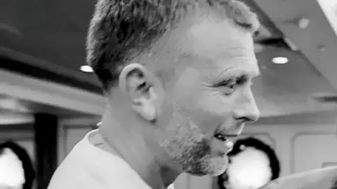 A black and white photo of a man supporting a young boxer in a ring, with a crowd watching.
