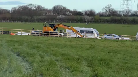 Supplied A green field with a barbed wire fence running through the middle. At the back of the field, there are several diggers roam around and piles of gravel are stacked up. 