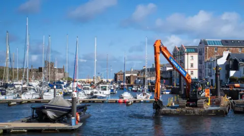 Manx Scenes Shot of Peel Marina on a sunny day. Sailing boats are docked in the water whilst the castle is seen on the left.