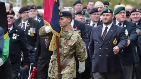 St Helens Council A young male soldier in Army cap and camouflage uniform jacket leads a march while holding a ceremonial standard flag. Veterans in dark suits, regimental caps and medals on lapels march behind him during the parade.