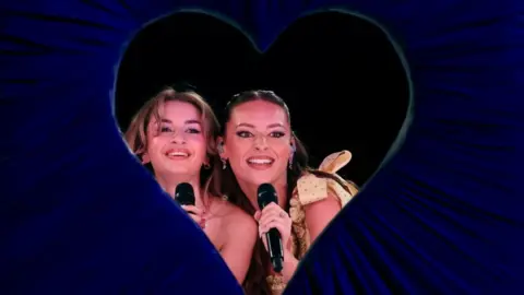 Reuters Two of the three members of girl band Remember Monday during a dress rehearsal, looking through a stage prop heart-shaped blue velvet curtain while holding microphones and smiling.