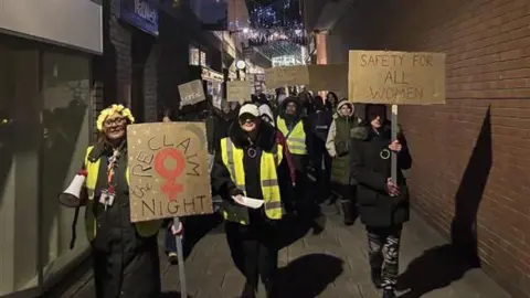 Restore A crowd of people with placards walking through a shopping centre ally. It is dark and some people are wearing yellow high-vis vests.