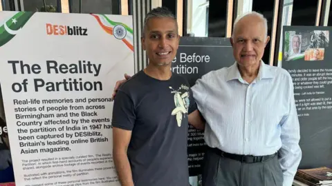 DESIblitz Two men stand in front of a banner titled: The Reality of Partition exhibition