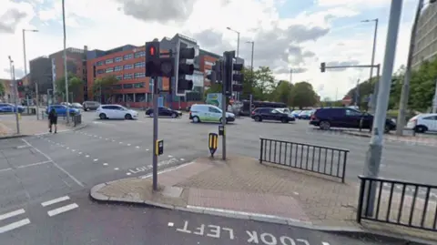 A wide road junction with traffic lights and pedestrian crossings and tall buildings on the far side