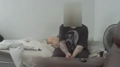 A man with a blurred face is sitting on a messy bed wearing a black T-Shirt.