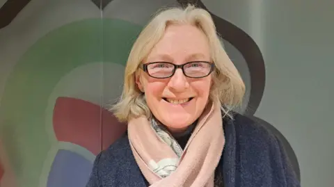 A woman with slightly shorter than shoulder length blonde hair stands wearing a grey coat and pink scarf against the backdrop of a West Northamptonshire Council logo.