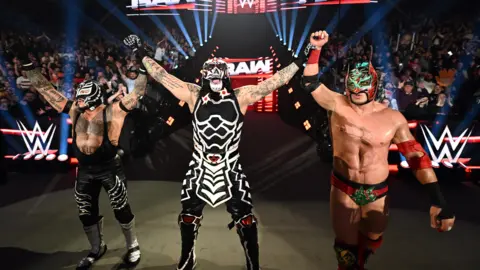Getty Images Three wrestlers are standing hand in hand. They are all in costume with masks on. 