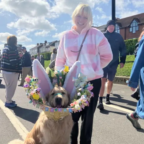 A woman stands on a residential street holding a large retreiver dog on a lead. The dog wears a colourful decorative headpiece with upright bunny ears and a floral wreath around its neck. Other people walk along the street behind them, with houses, grassy verges, and a bright, partly cloudy sky visible in the background.