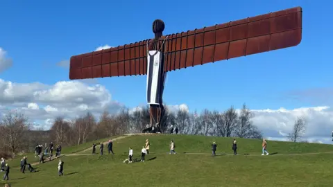 BBC The Angel of the North dengan kemeja Newcastle United hitam dan putih tergantung dari tubuhnya. Orang dapat dilihat di dasar bukit.