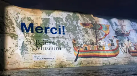 Stâncile albe din Dover, la Muzeul Britanic, noaptea, acoperite de o proiecție gigantică cu imagini cu bărci de lemn din tapiserie și „Merci”