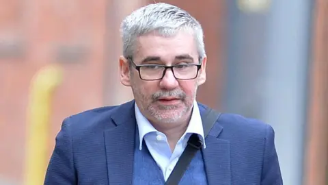A man with short grey hair, wearing black rectangular glasses, a blue blazer, a blue waistcoat and a shirt opened at the collar