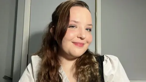 Aeryn Hill smiles at the camera and looks to the side. She has long wavy brown hair and is wearing white