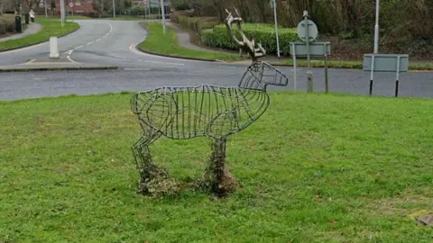 The wire-framed stag sculpture in the middle of a grass-covered roundabout with a road snaking of in the background