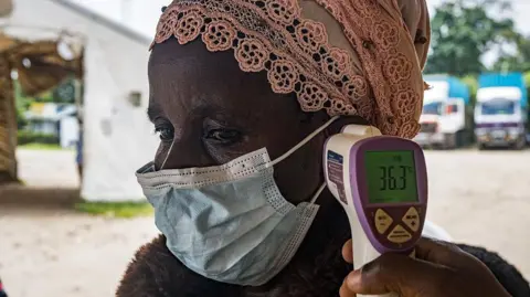 A person wearing a mask and peach headwrap is having their temperature checked with a contactless thermometer near their ear. it reads 36.3 degrees.