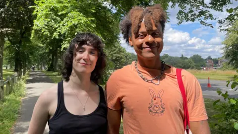 Sofía Luis-Hobbs/BBC A brunette person with shoulder length wavy hair smiles at the camera wearing a black tank top. To their right, a person with curly short hair smiles at the camera wearing an orange tshirt with a bunny graphic on the front. They have a silver chain with a heart on it. They are both on a footpath surrounded by greenery on a bright day.