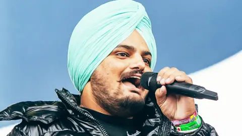 Getty Images Moose Wala is wearing a black puffa jacket, turquoise turban and is singing into a microphone.